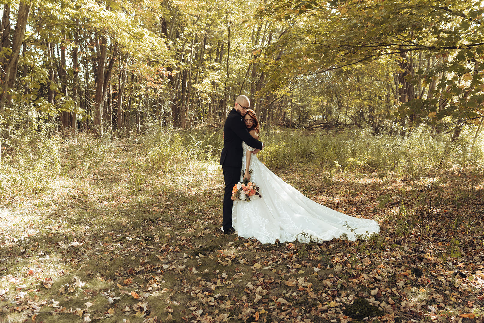 A bride and groom kiss in a sunlit autumn forest during fall wedding portraits, the bride's lace gown and train spread across golden leaves — Hudson Valley wedding photography by Rob Charles Photography.