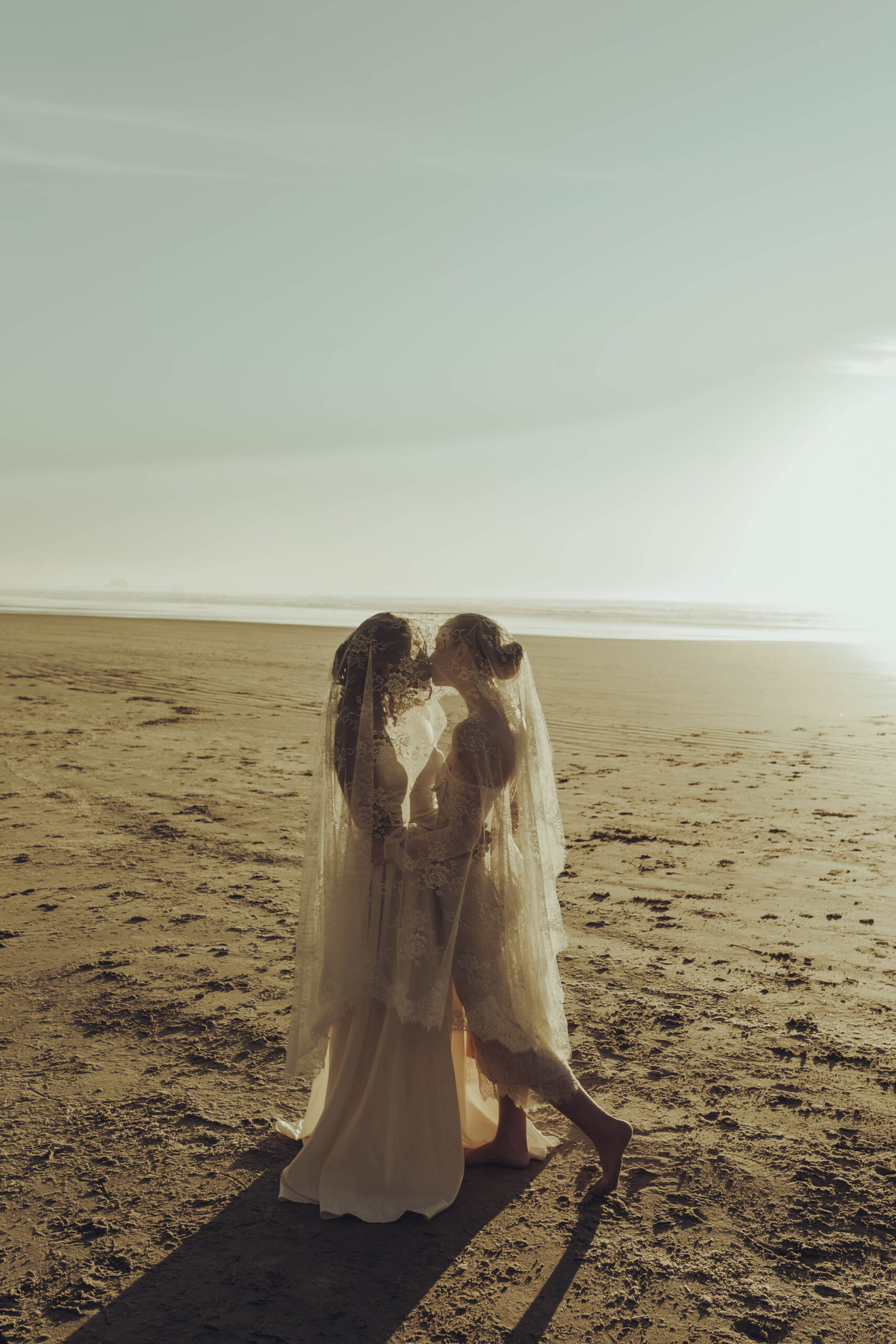 Two brides share a romantic kiss beneath their lace veils on a sun-drenched beach during their elopement — LGBTQIA+ inclusive wedding photography by Rob Charles Photography.